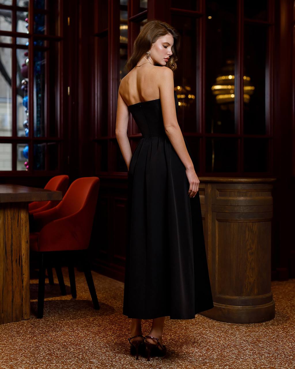 Woman in a black dress standing in a dimly lit room with wooden furniture.