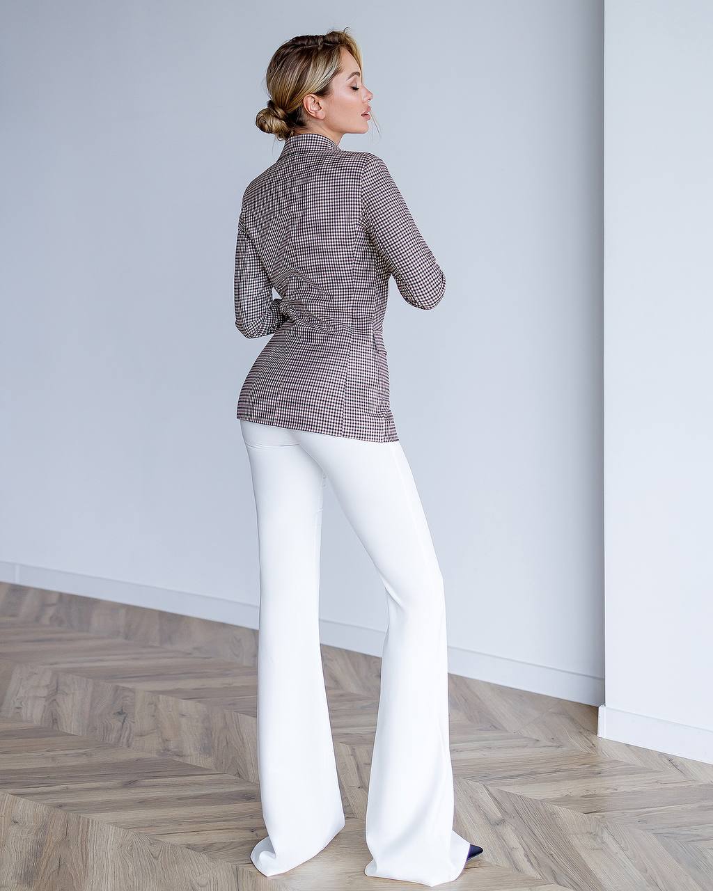 Woman wearing a patterned blazer and white pants standing against a plain wall.