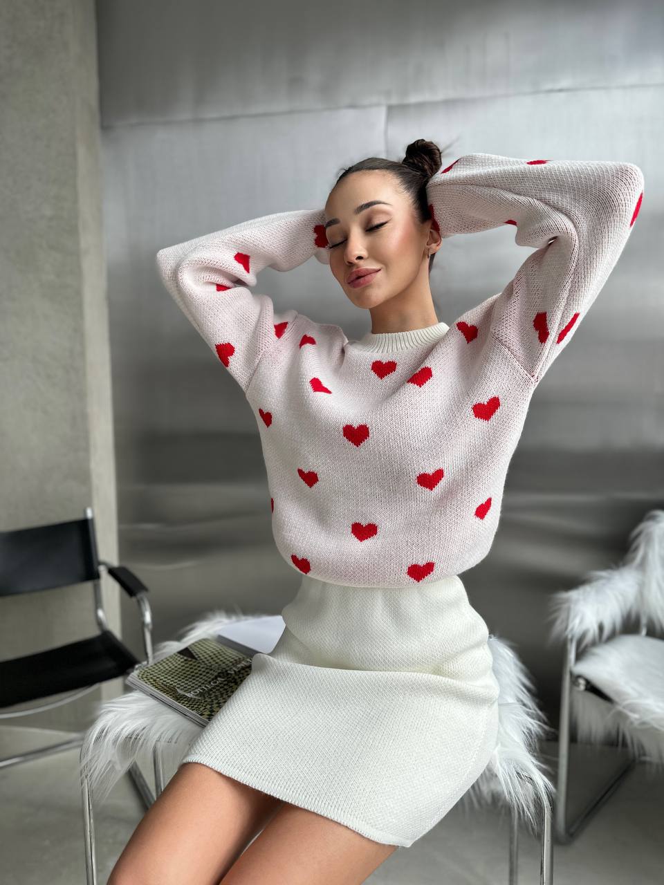 Woman wearing a white sweater with red heart patterns and a white skirt in a modern room.