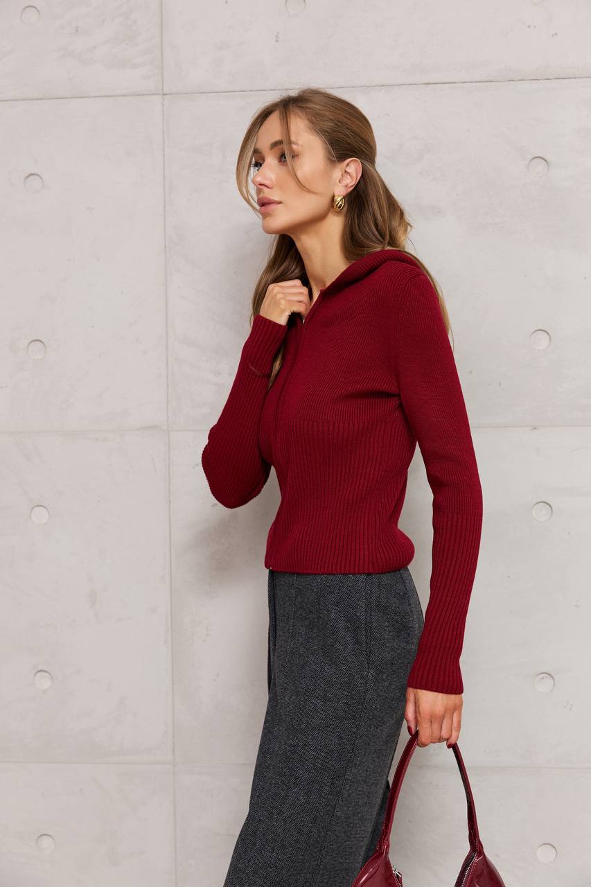 Woman wearing a red sweater and gray pants against a light gray wall.