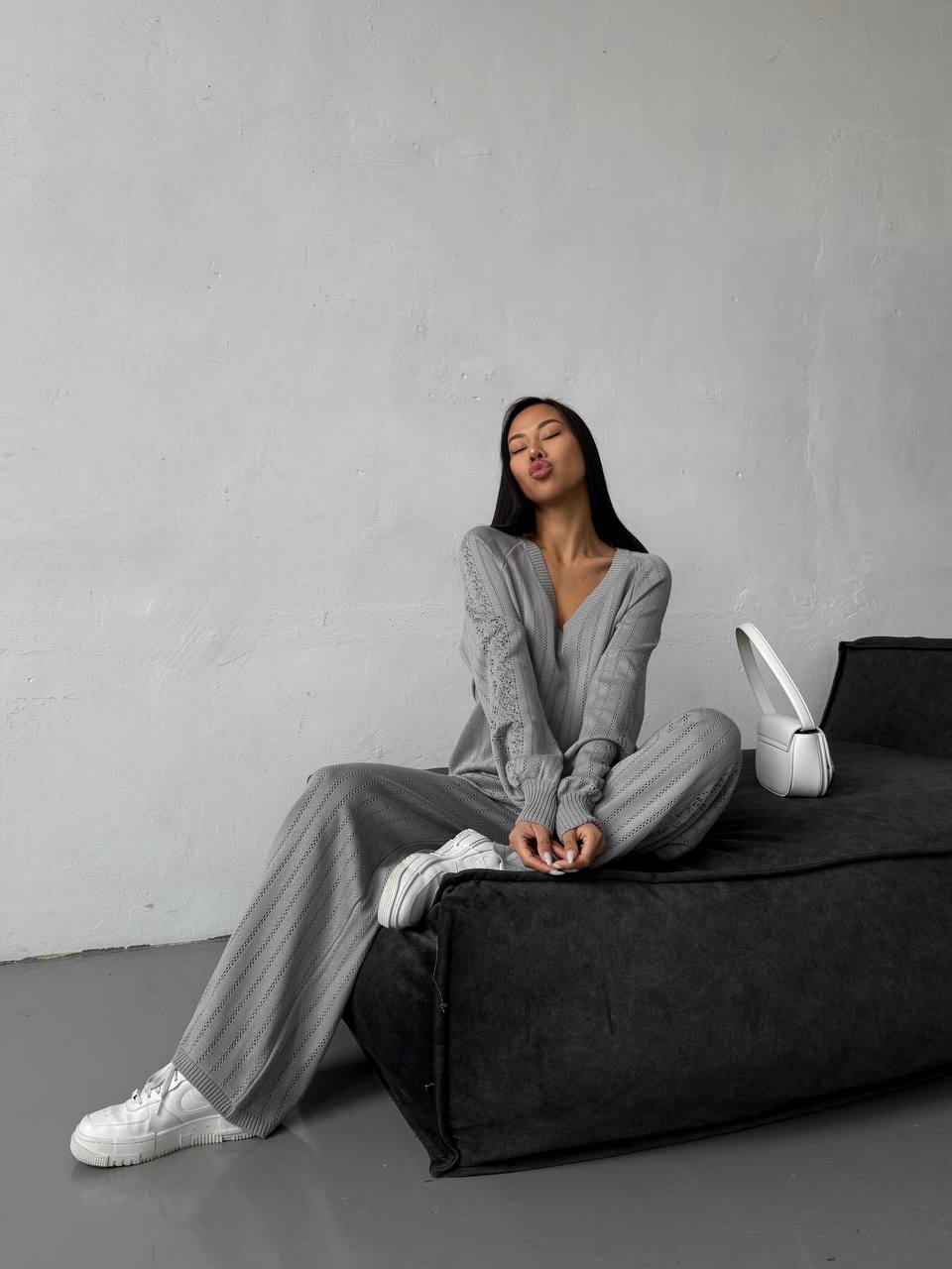 Woman in gray outfit sitting on a black bean bag chair against a plain wall.