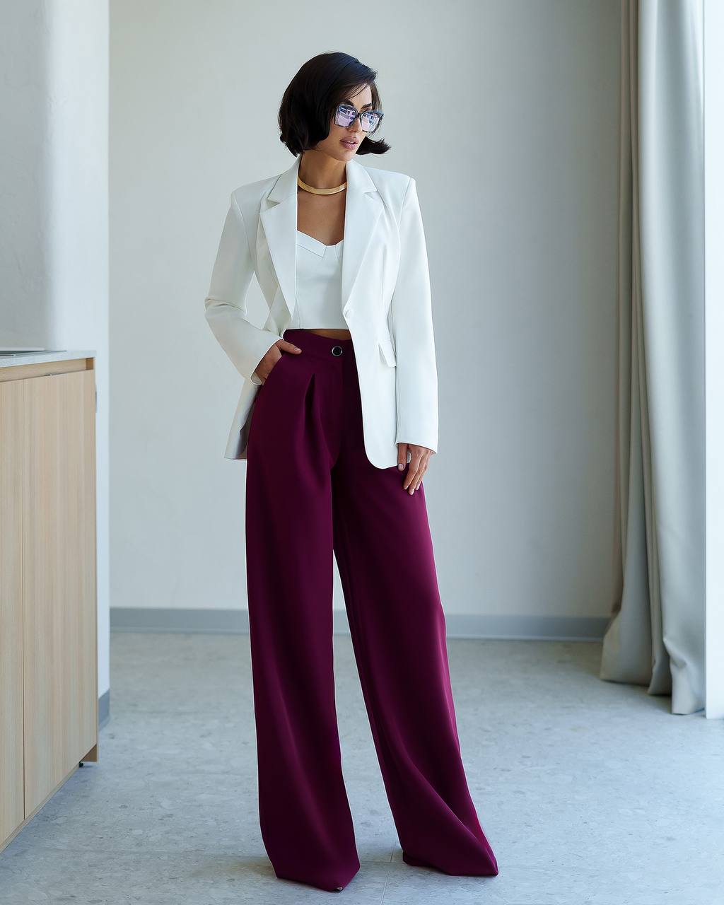 Woman wearing a white blazer and burgundy pants in a minimalistic room.
