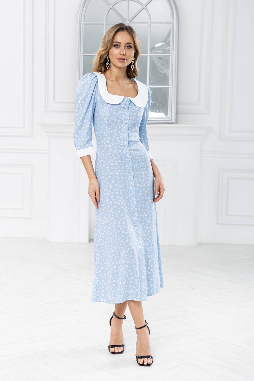 Woman wearing a light blue dress with white collar in a white room.