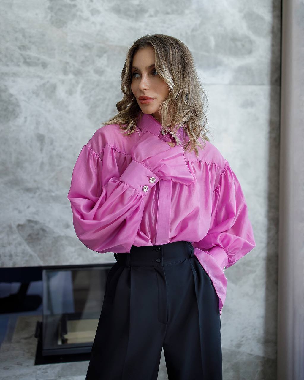 Woman wearing a bright pink shirt and black pants against a gray wall.