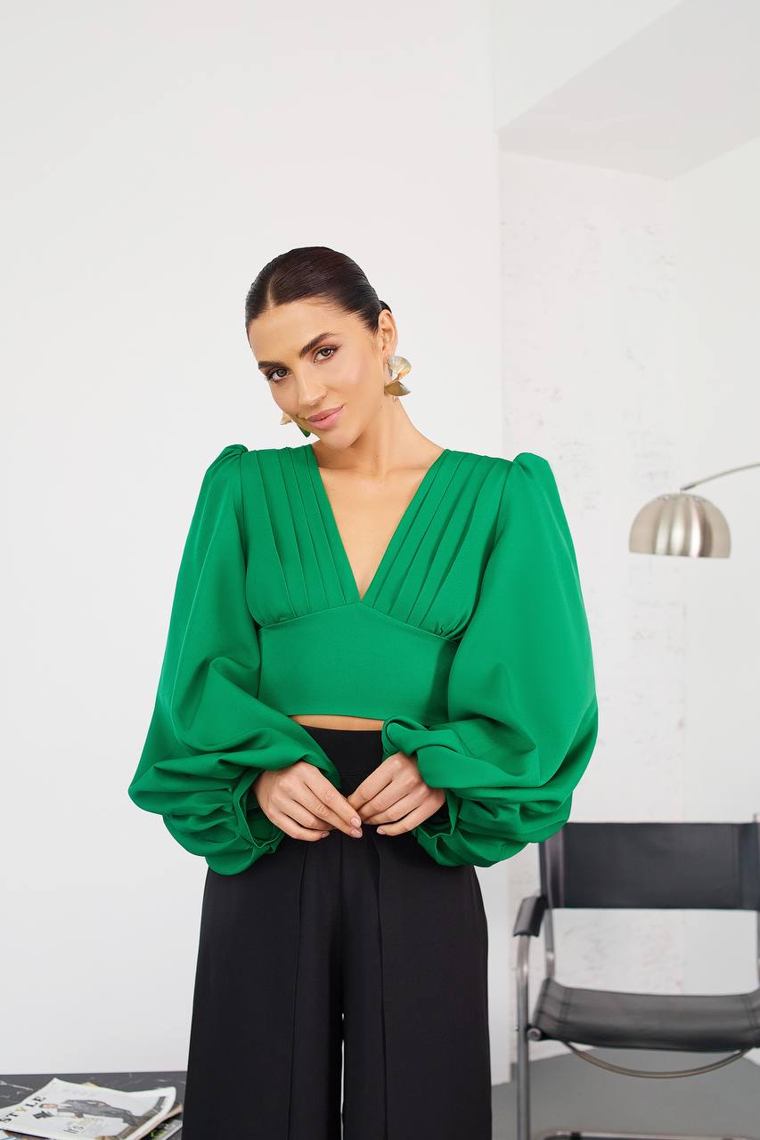 Woman wearing a green blouse with puffed sleeves and black pants in an indoor setting.