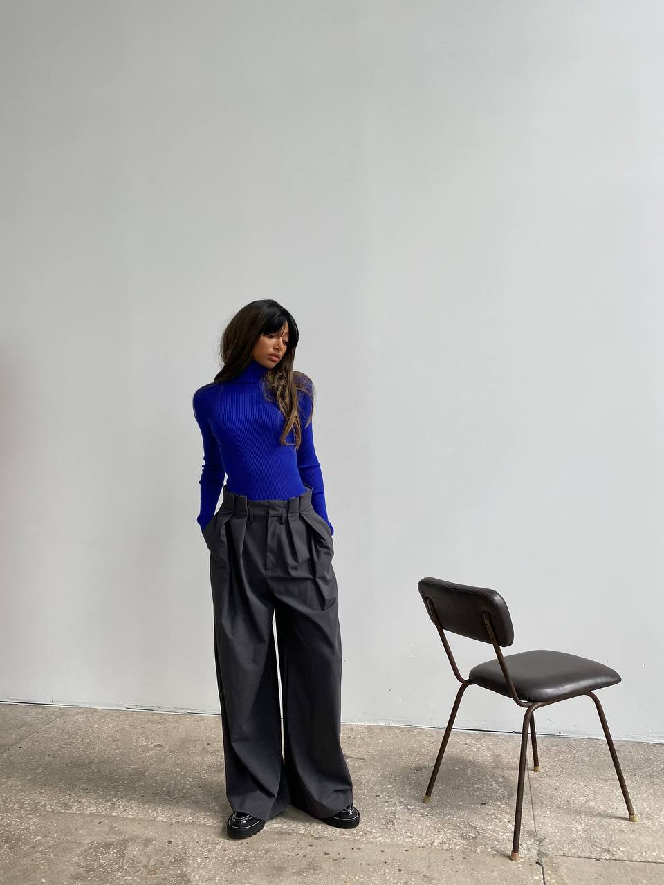 Woman in a blue top and black pants standing next to a chair against a plain wall.
