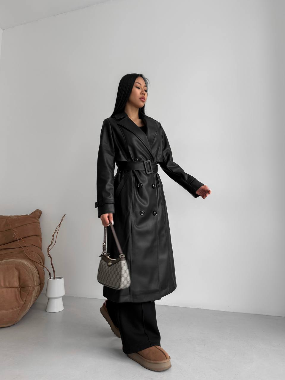 Person wearing a black trench coat holding a handbag in a minimalistic setting.