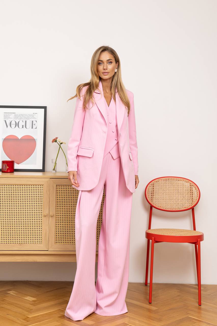 Woman in a pink suit standing in a room with a wooden cabinet and red chair.