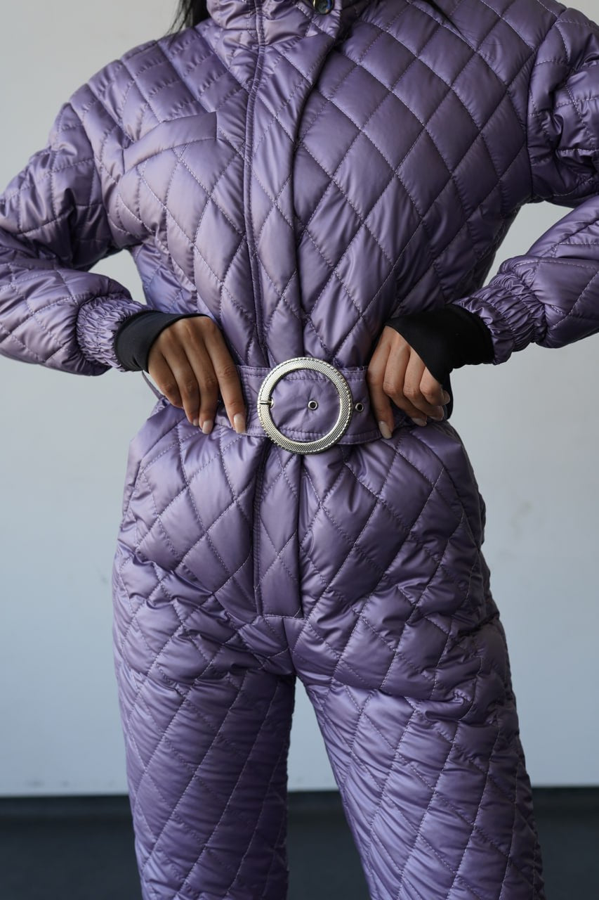Person wearing a purple quilted jumpsuit with a belt against a neutral background