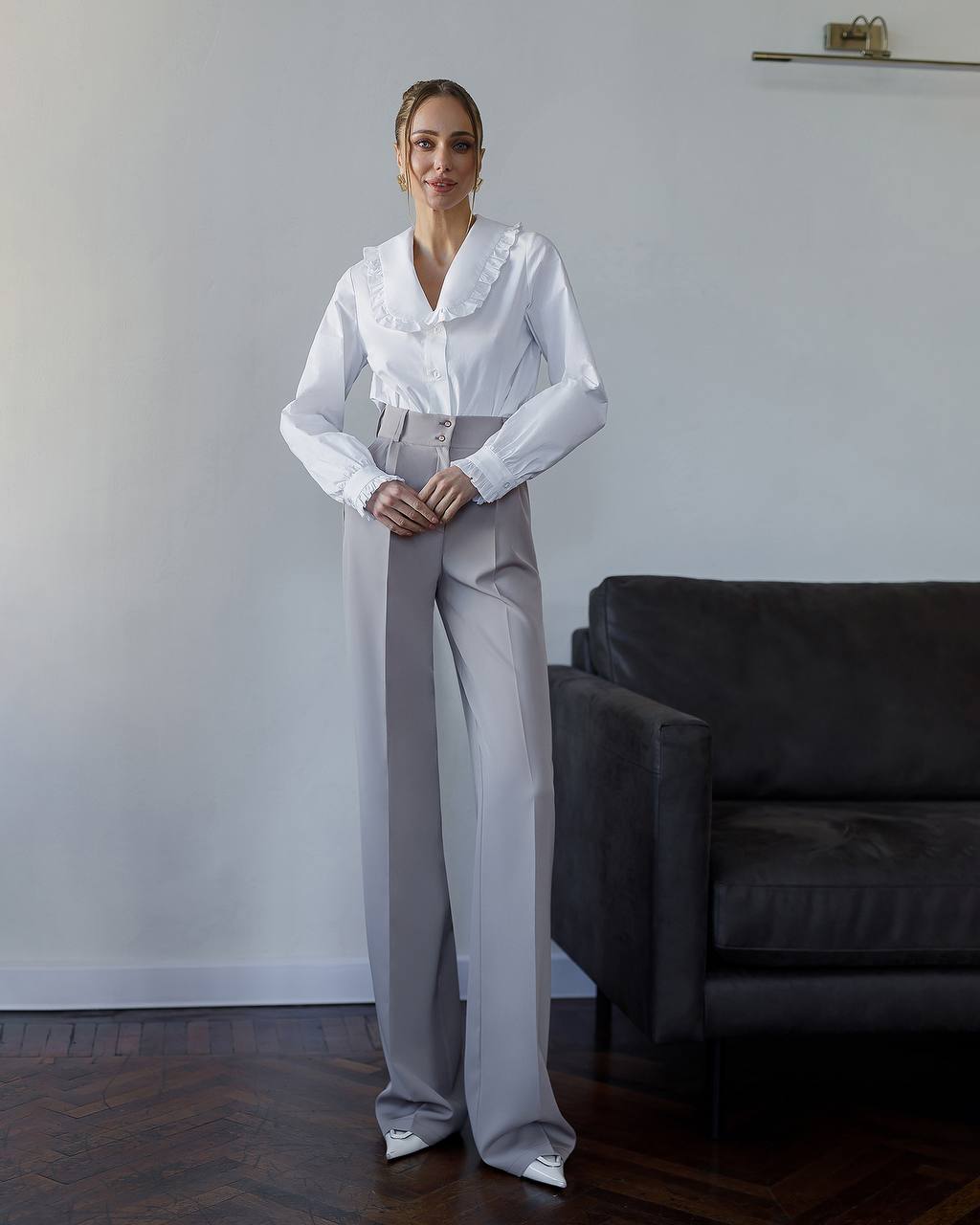 Woman in a white blouse and gray pants standing in a room with a dark sofa.