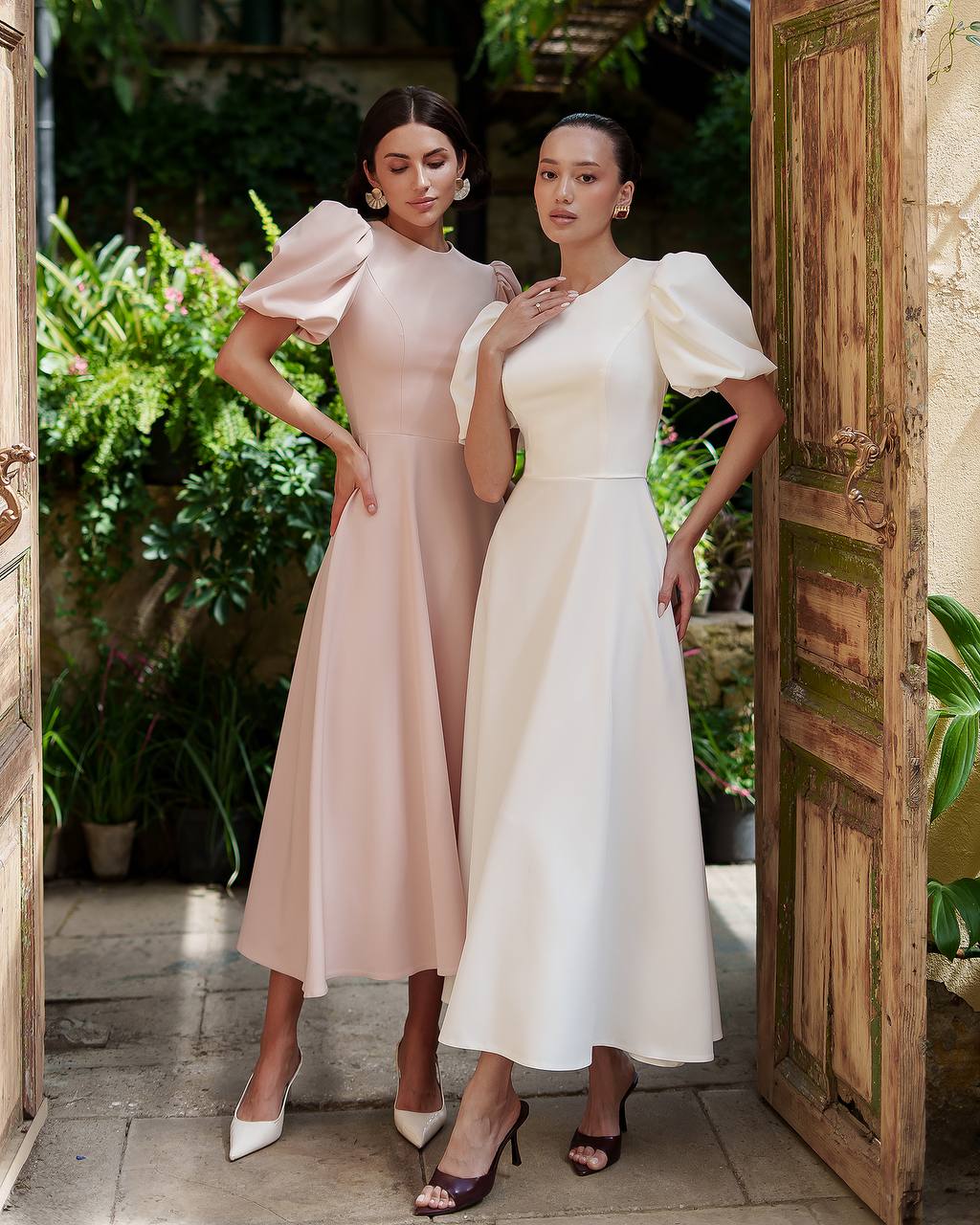 Two women in elegant dresses standing in a doorway with greenery in the background