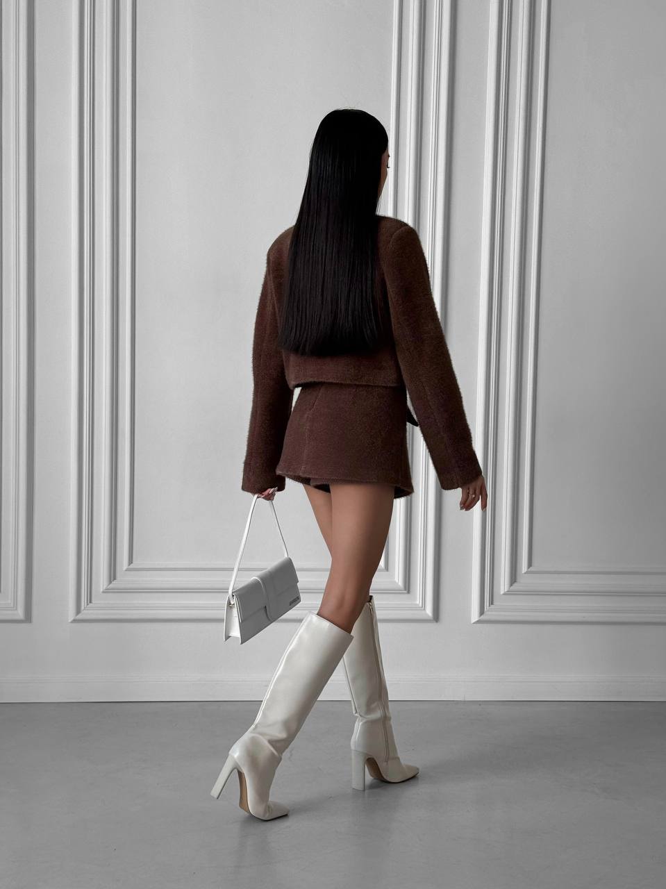 Woman in a brown outfit with white knee-high boots walking away against a gray wall.