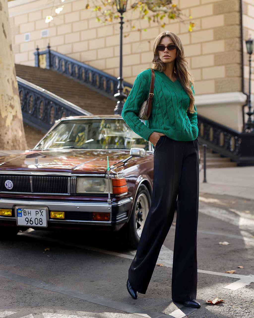 Woman in a green sweater and black pants standing next to a vintage car on a city street.