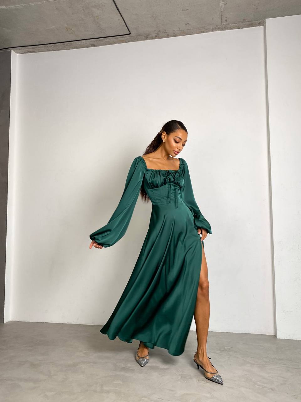 Woman wearing a green dress with long sleeves and a slit, standing against a white wall.