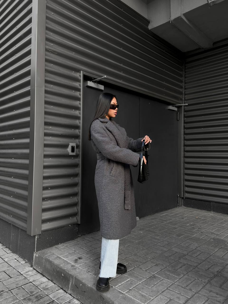 Person in a long coat standing outside a building with corrugated metal walls.