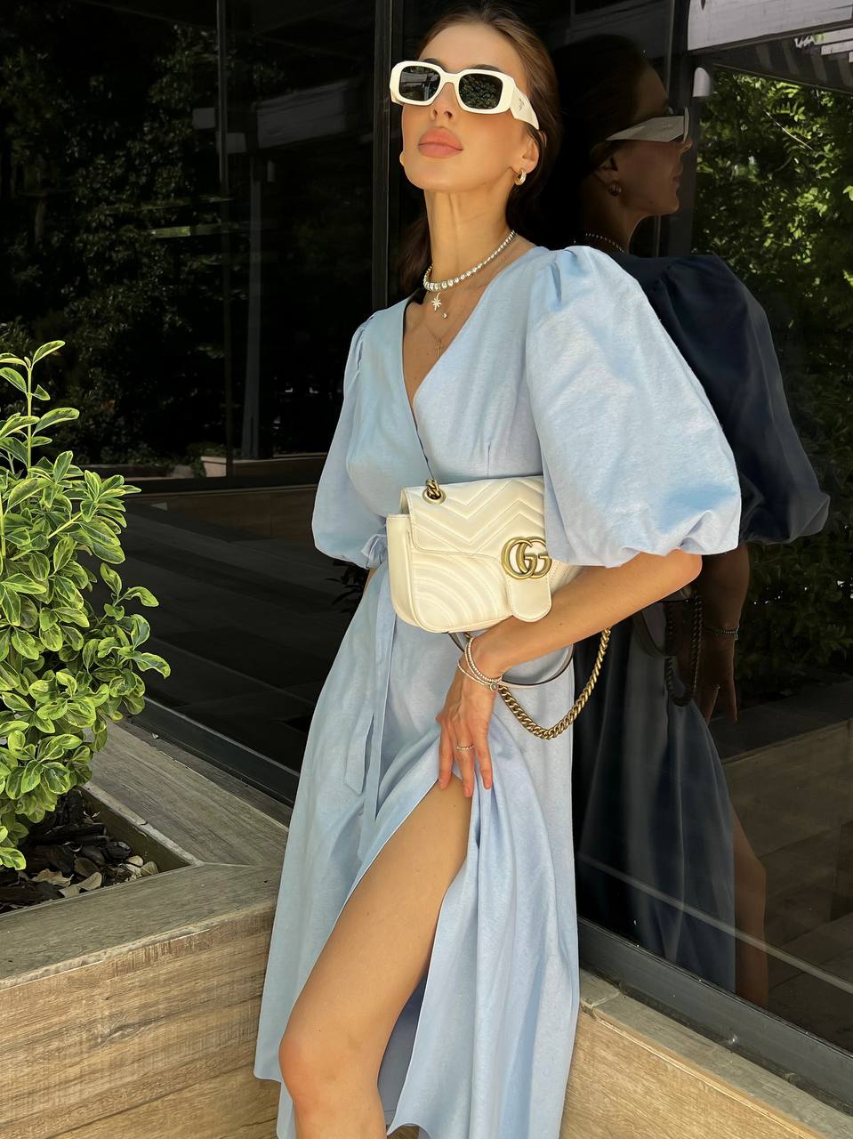 Woman in a light blue dress with a white handbag, standing outdoors.