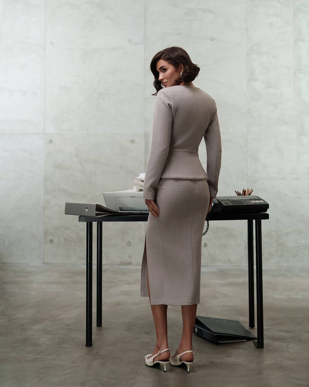 Woman in a gray suit standing in front of a minimalist desk with a concrete wall background
