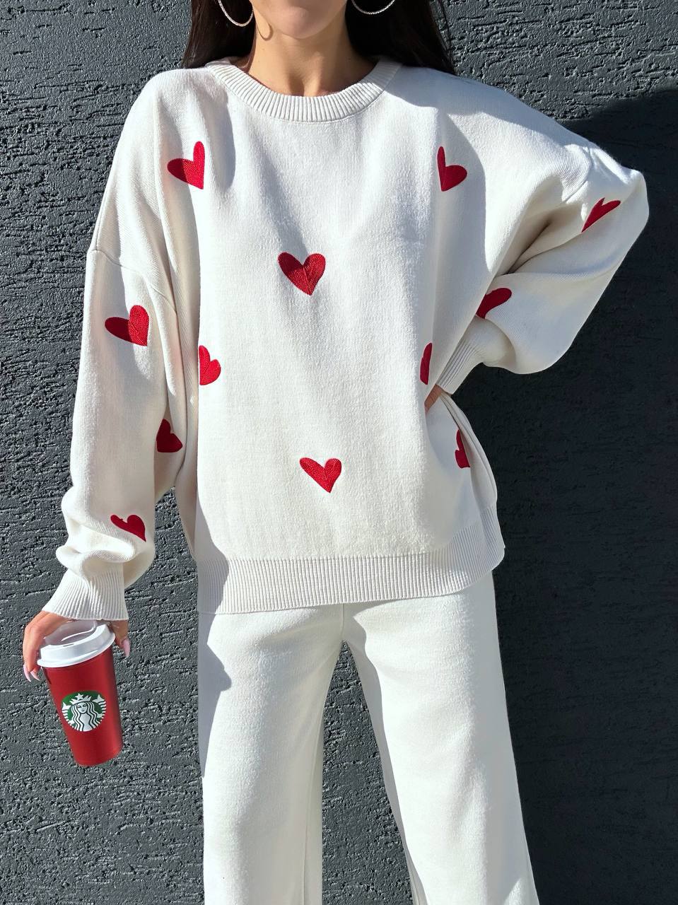 Person wearing a white sweater with red heart patterns against a gray wall.