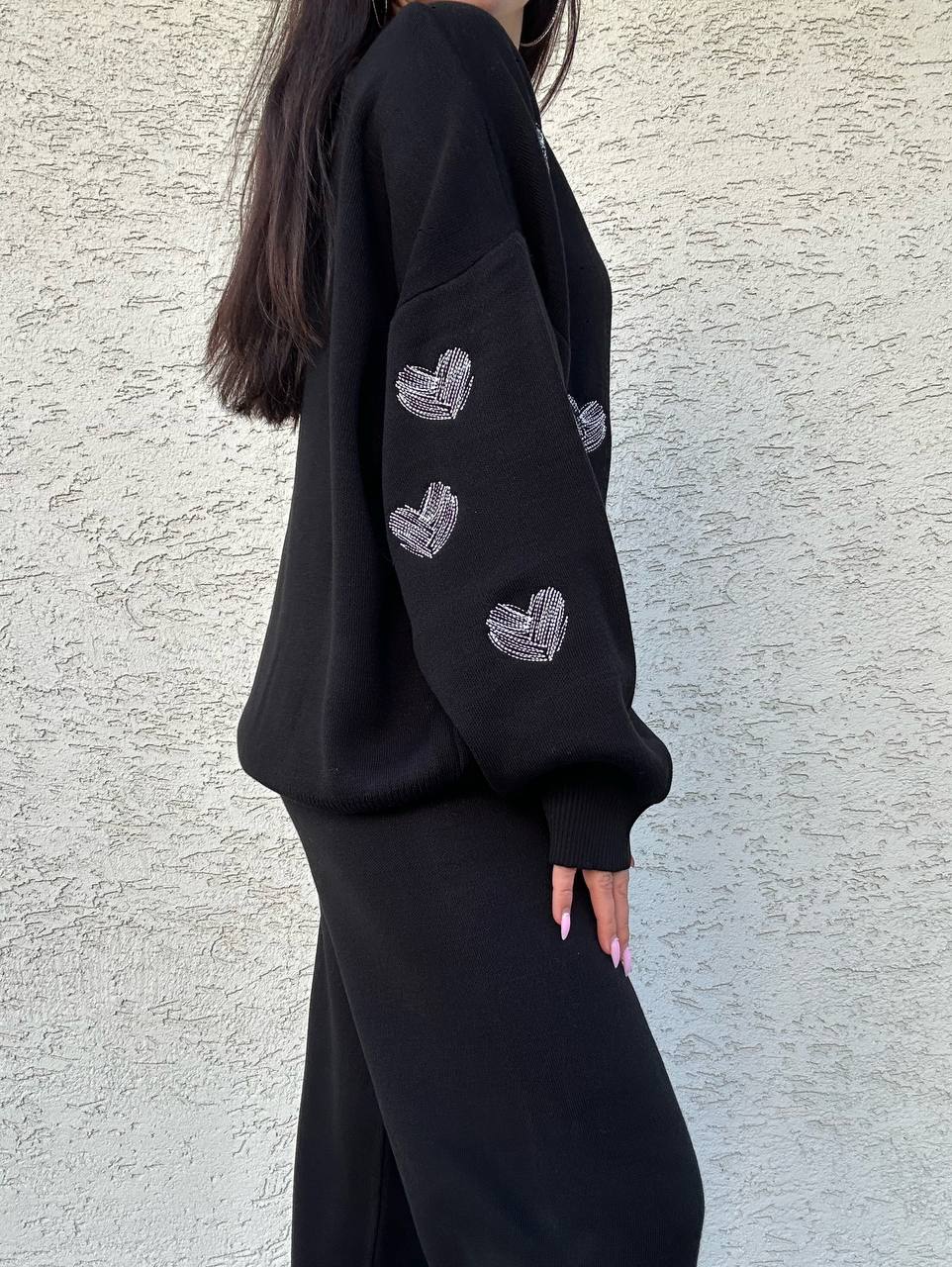 Person wearing a black sweatshirt with heart patterns on a light gray wall.