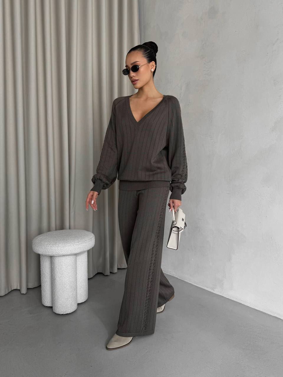 Woman in a gray outfit standing in a minimalistic room with a curtain and stool.