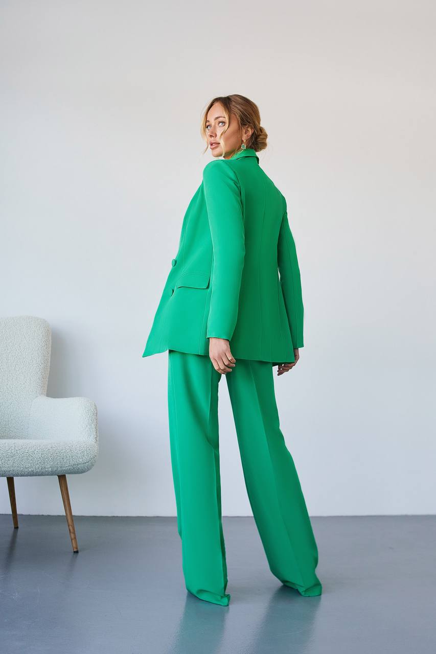 Person wearing a green suit standing against a white wall with a light gray chair.
