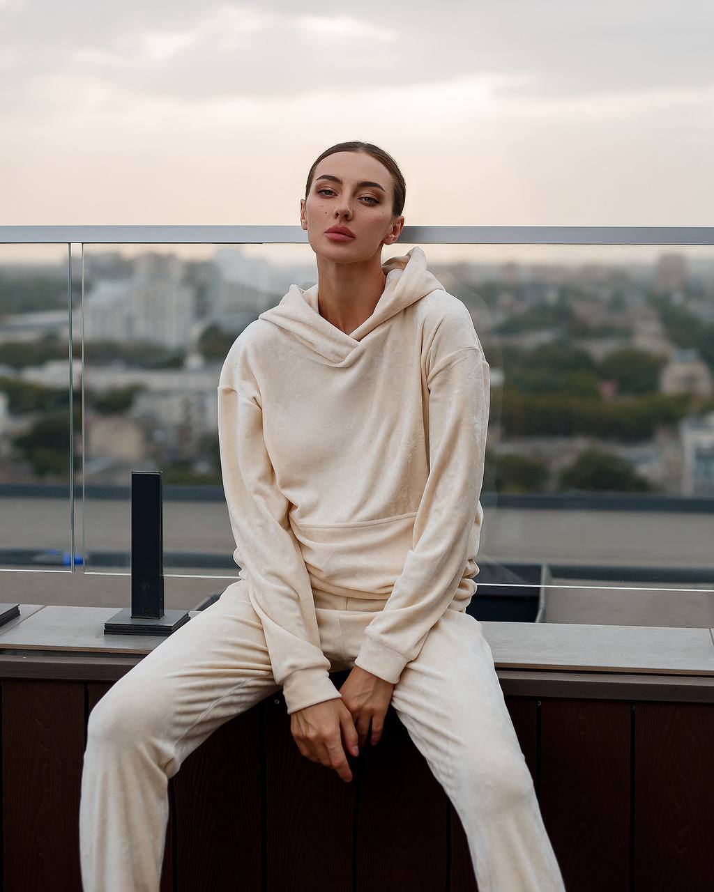 Person wearing a cream-colored hoodie and pants sitting on a ledge with a cityscape in the background