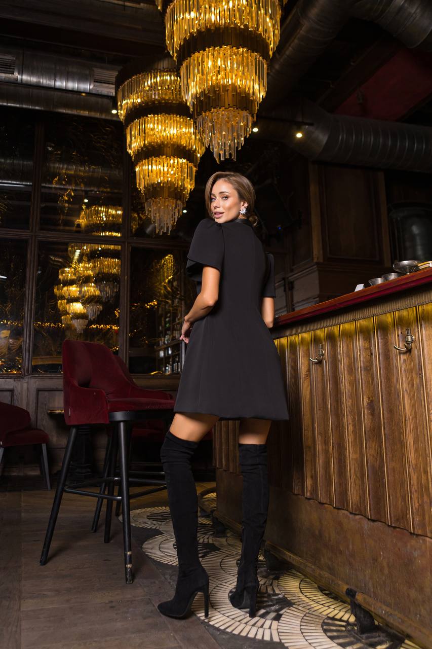 Woman in a black dress and knee-high boots standing in a stylish bar with chandeliers.