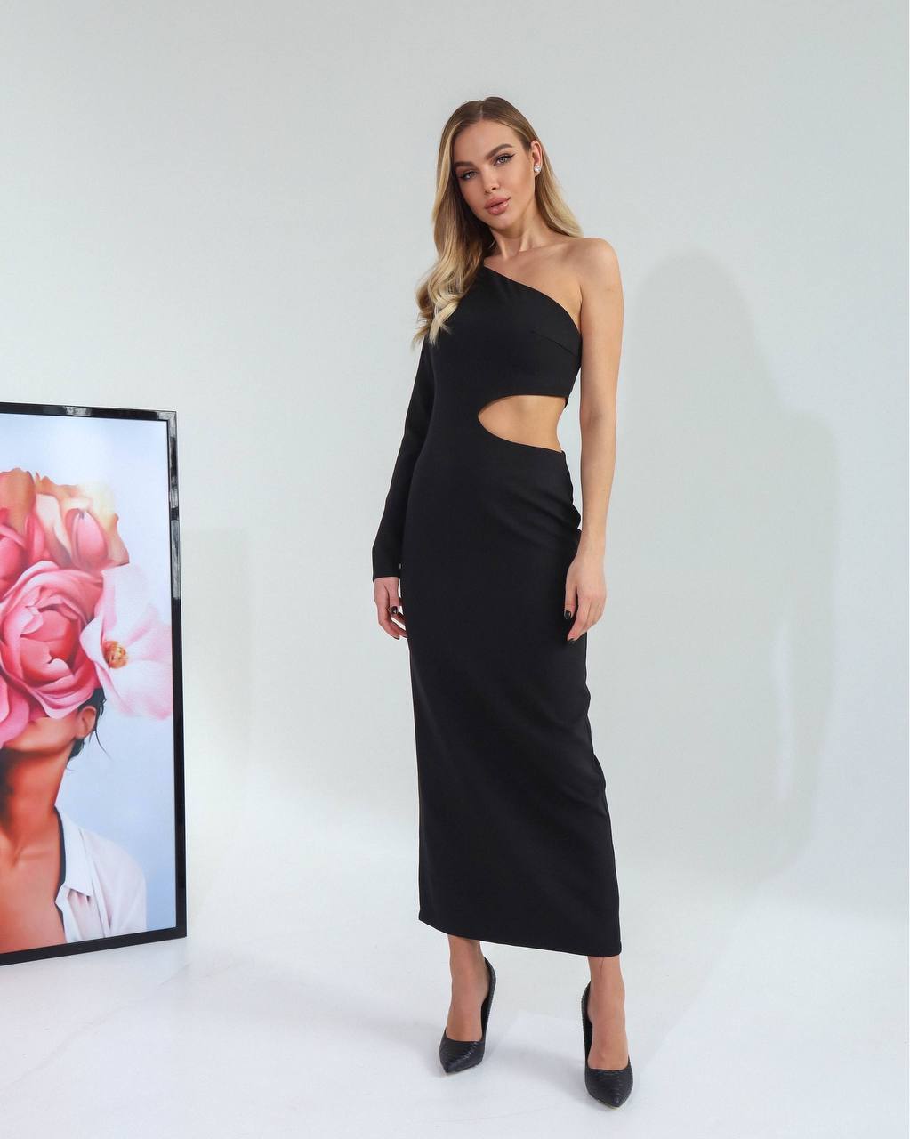 Woman wearing a black one-shoulder dress with a cutout design, standing against a white background.