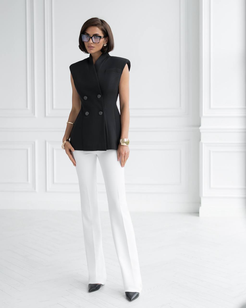 Woman wearing a black sleeveless blazer and white pants against a white wall.