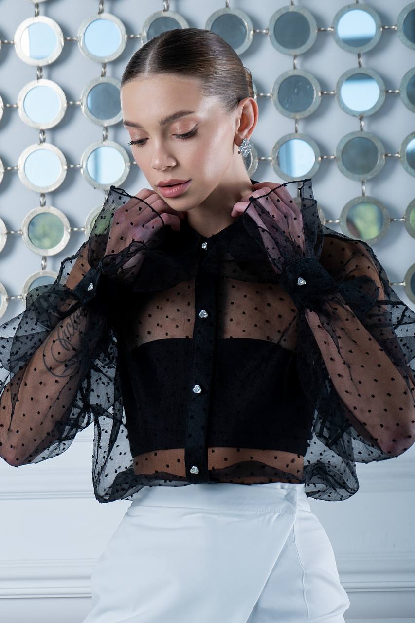 Woman wearing a black sheer blouse with white buttons against a decorative wall.