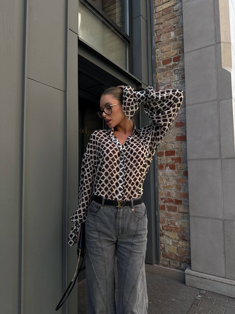 Person wearing a patterned shirt and jeans standing against a building.