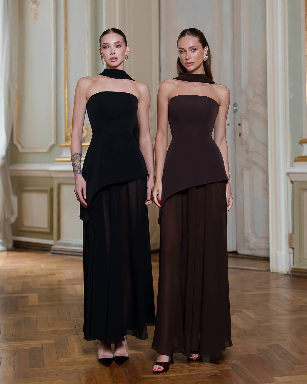 Two women wearing strapless black evening gowns in an elegant interior setting.
