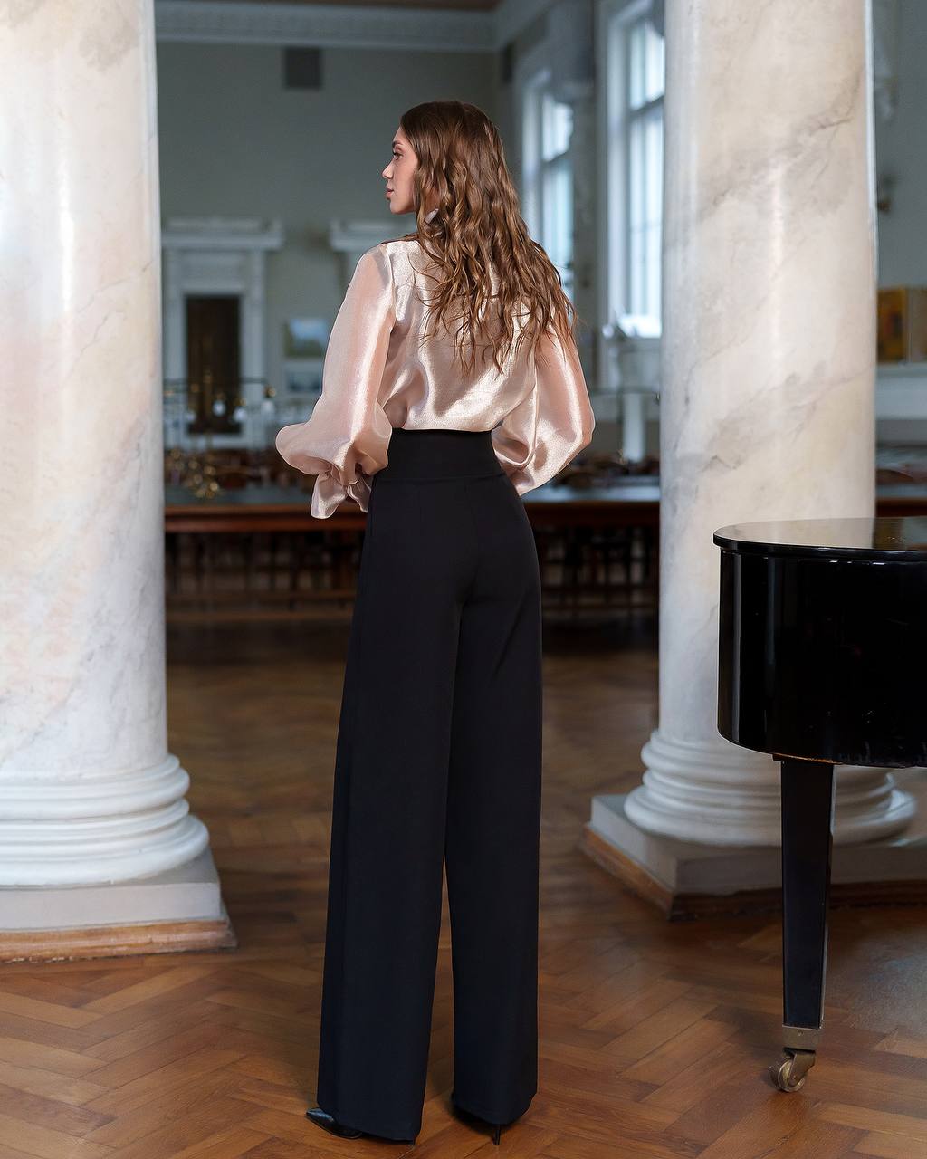 Woman in a beige blouse and black pants standing in a room with marble columns.