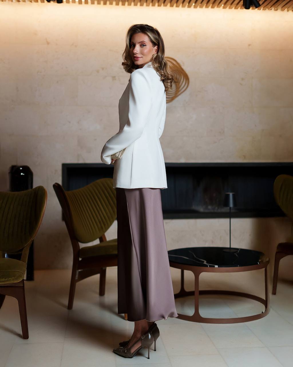 Woman in a white blazer and purple skirt standing in a stylish room with modern decor.