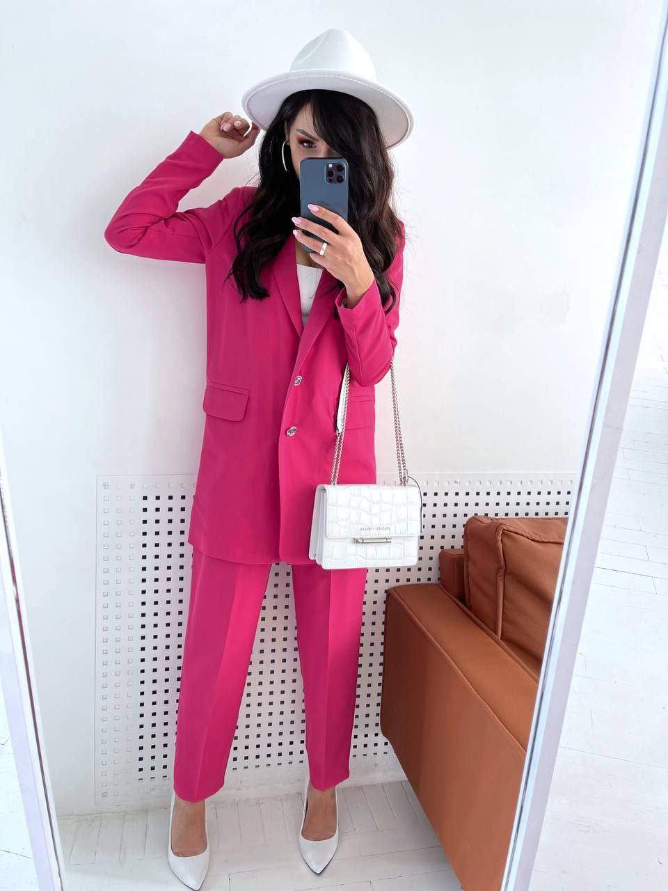 Person wearing a pink suit with a white hat and bag, taking a mirror selfie.
