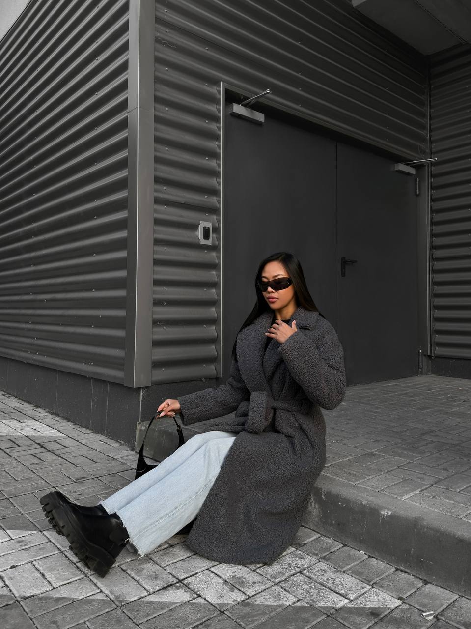 Person wearing a long coat sitting against a dark wall.