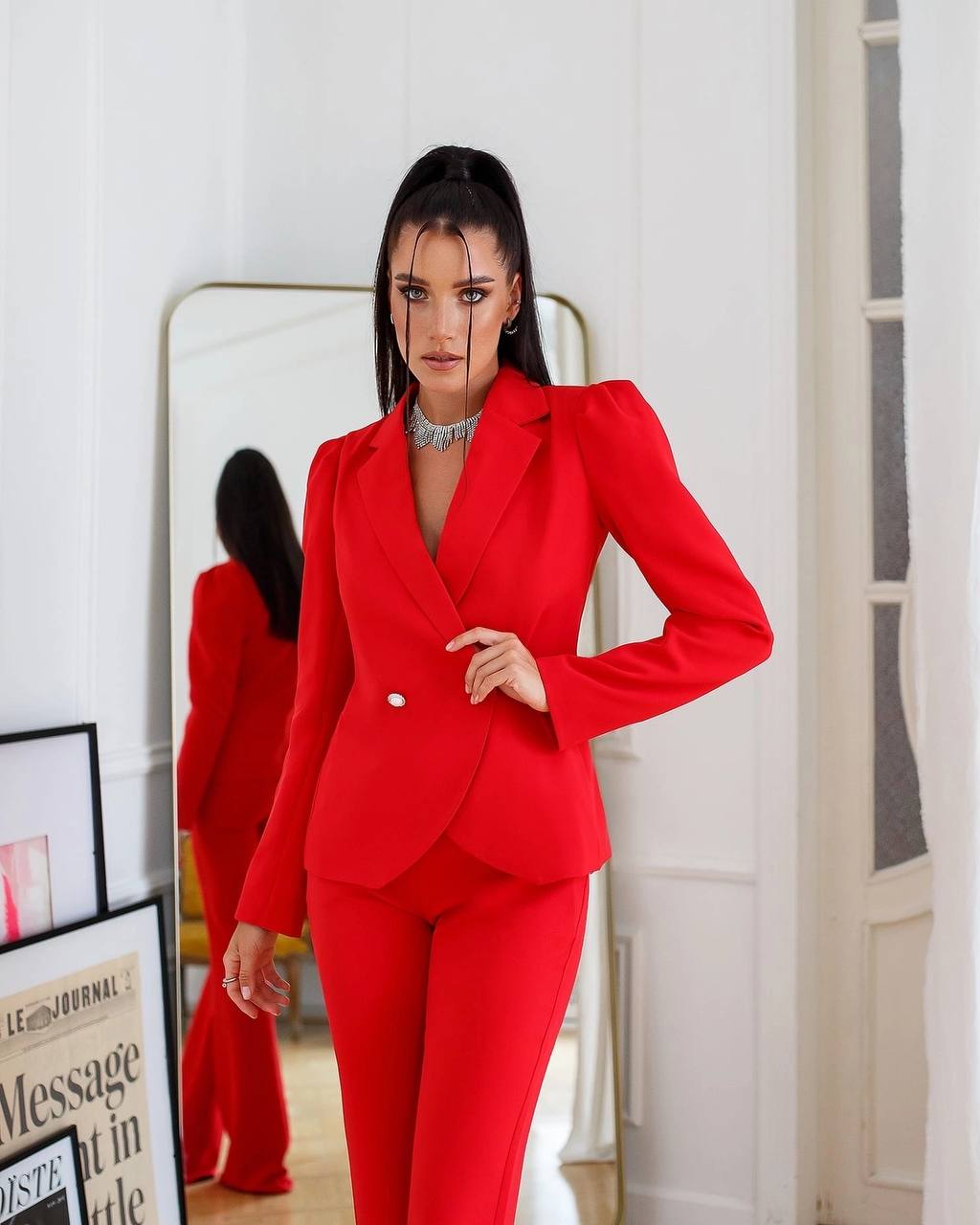 Woman wearing a red suit standing in a room with a mirror and framed picture.