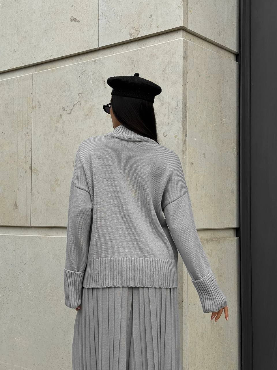 Person wearing a gray sweater and black beret against a stone wall.