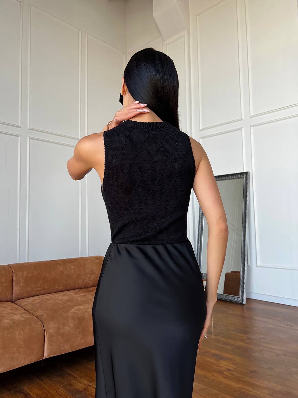 Woman wearing a black sleeveless dress standing in a room with white paneled walls and a brown sofa.