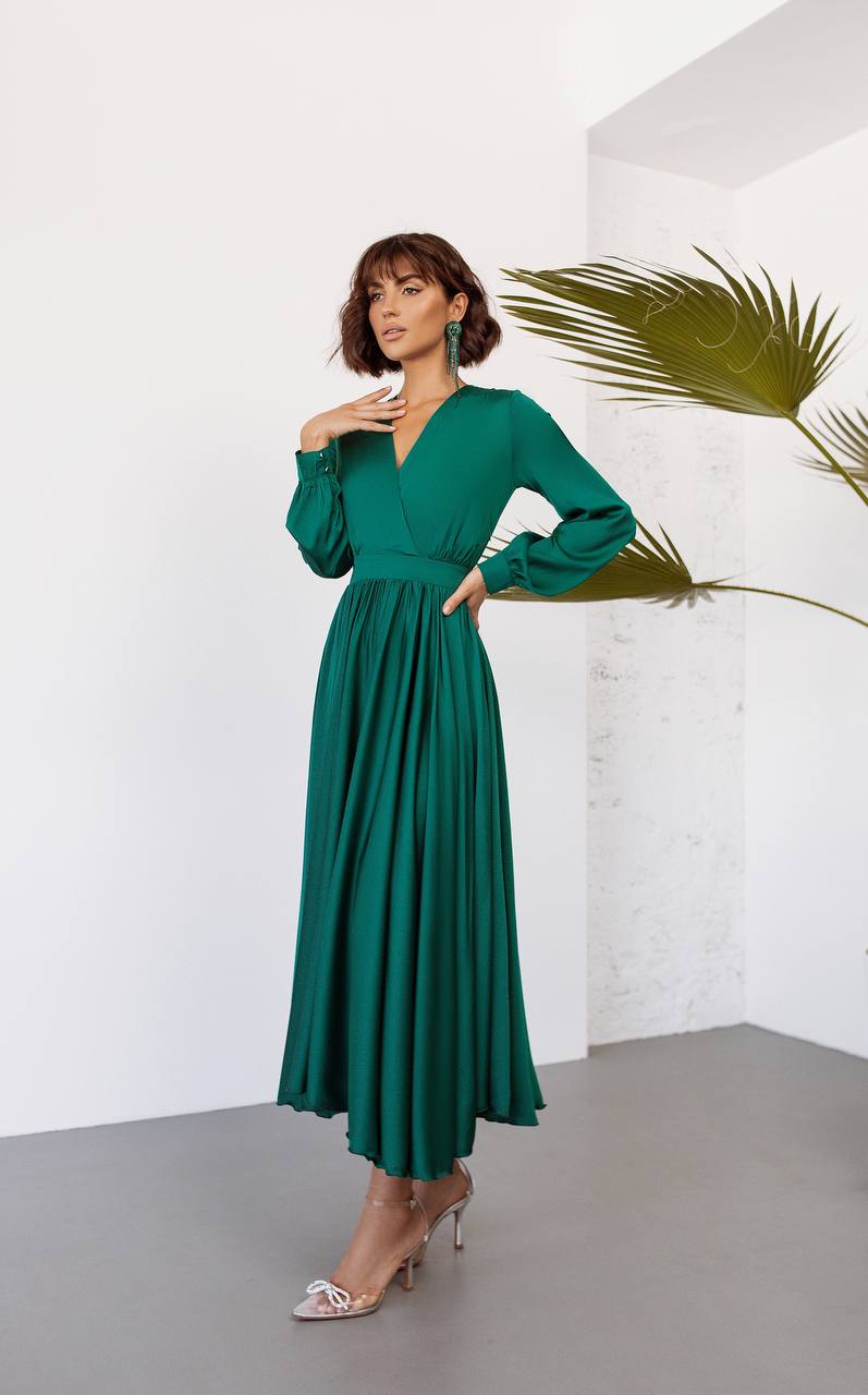Woman wearing a green dress standing against a white wall with a plant.