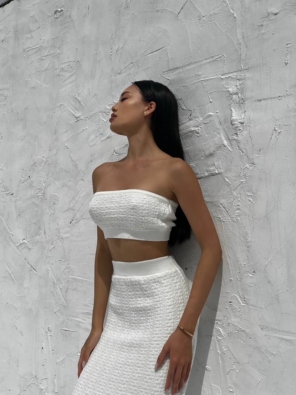 Woman wearing a white strapless top and skirt against a textured gray wall.