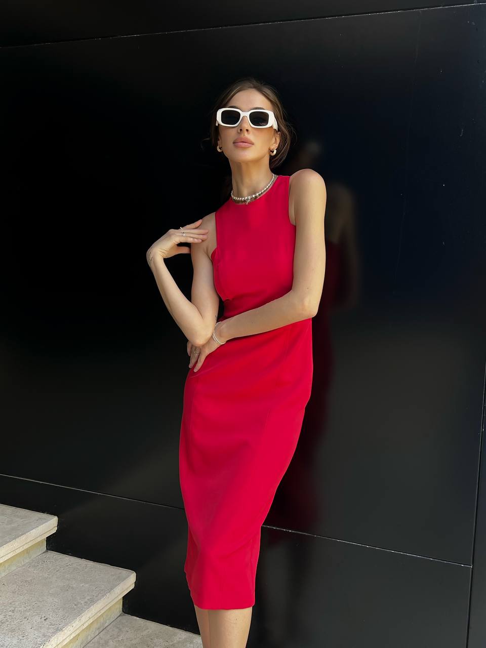 Woman in a red dress and sunglasses posing against a black wall.