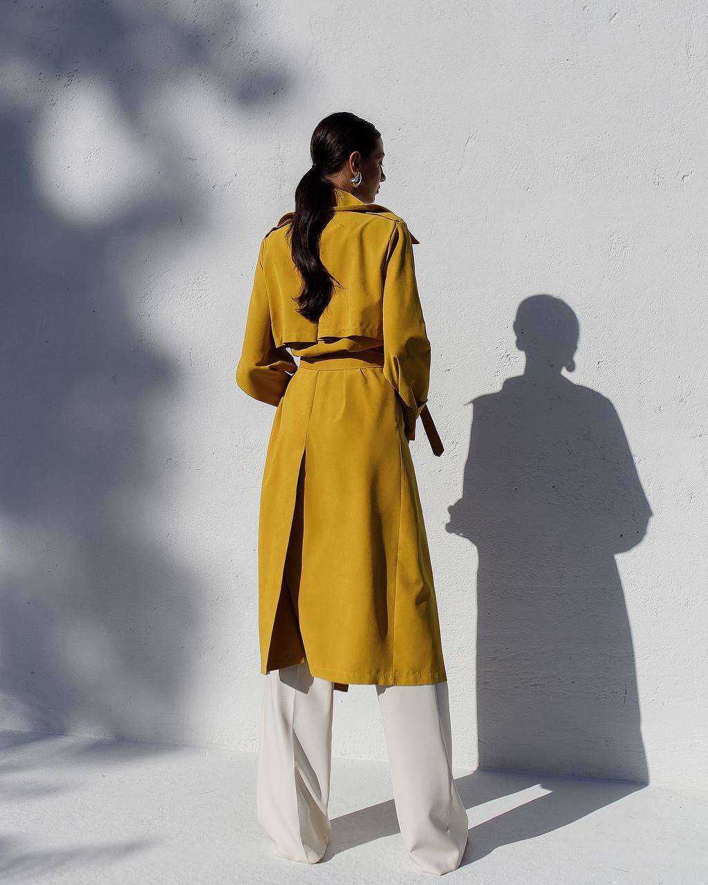Person wearing a yellow trench coat standing against a white wall with a shadow.