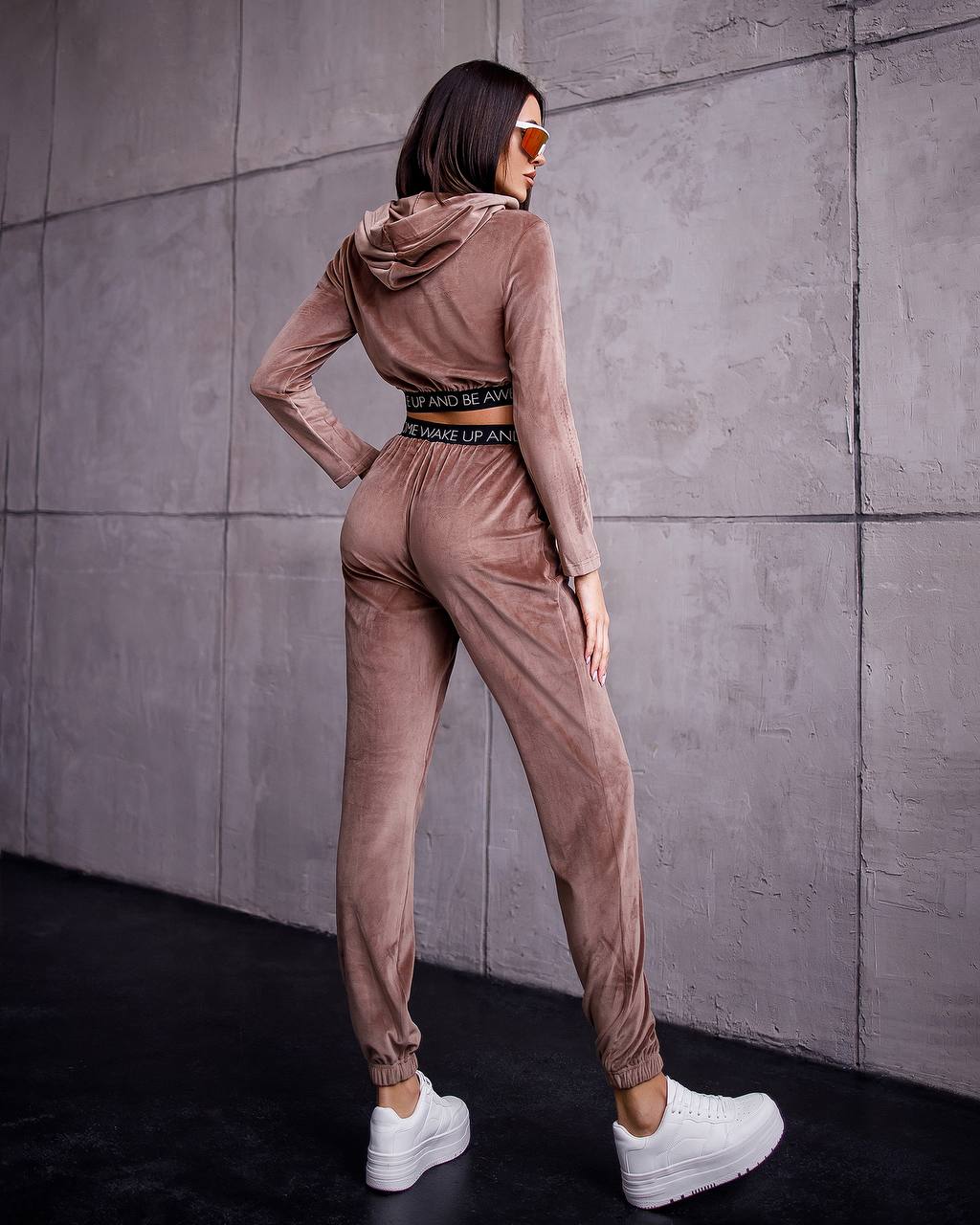 Woman wearing a brown tracksuit standing against a gray wall.