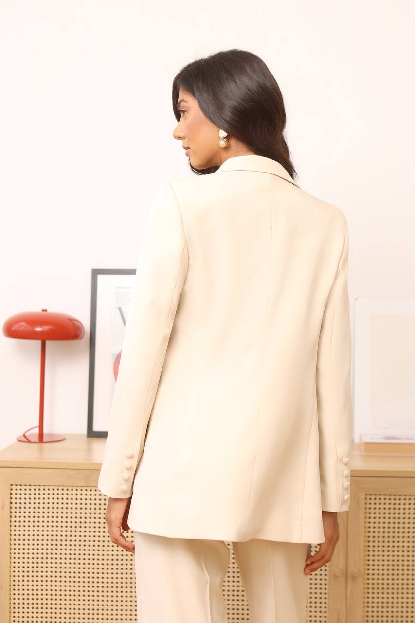 Woman wearing a beige suit standing in a room with a lamp and framed picture on a shelf.