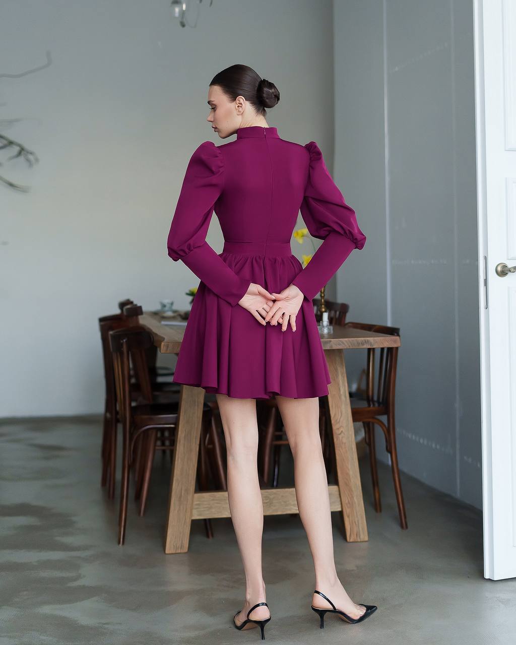 Woman in a purple dress standing in a room with wooden furniture.