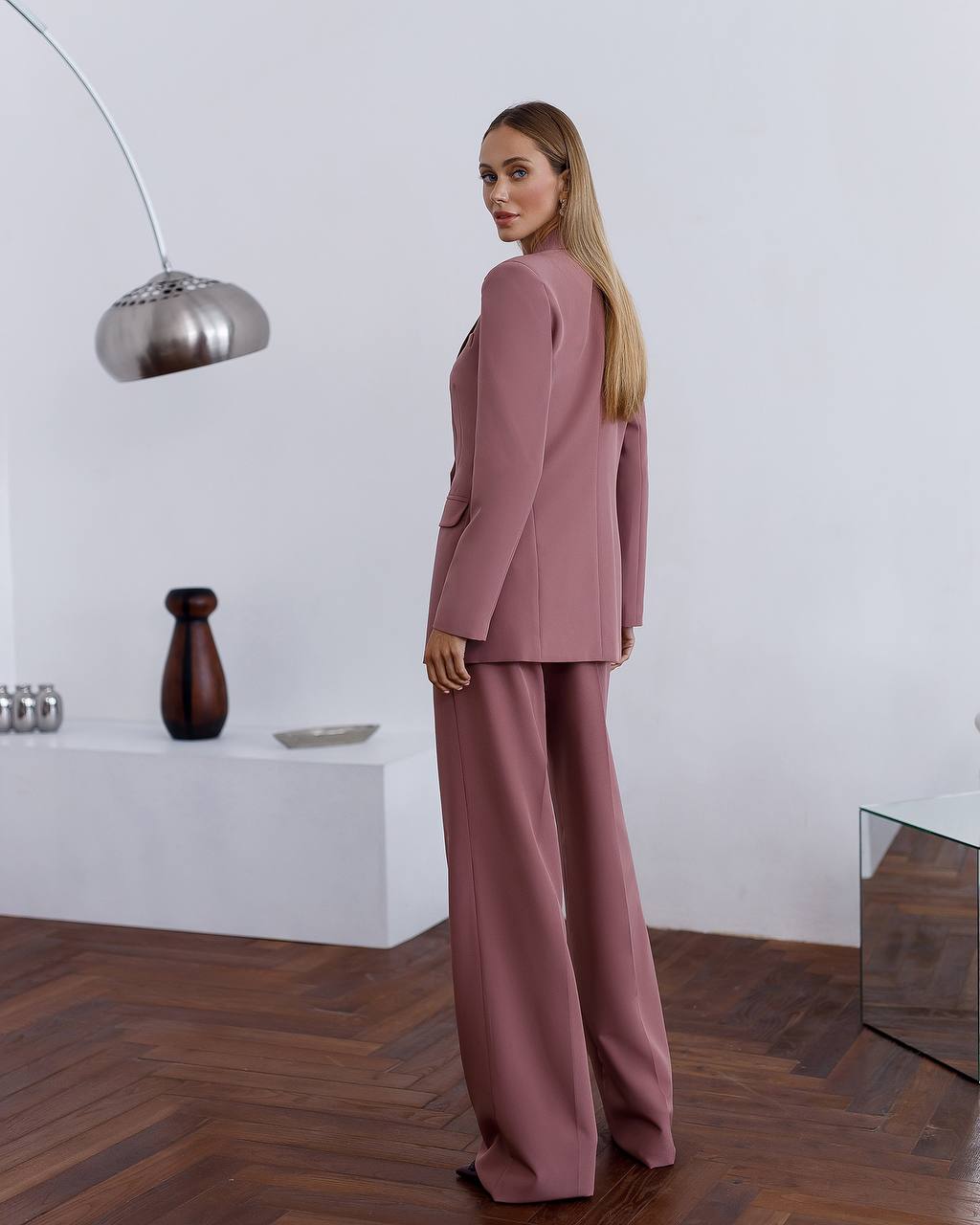 Woman in a pink suit standing in a modern room with wooden flooring and white walls.