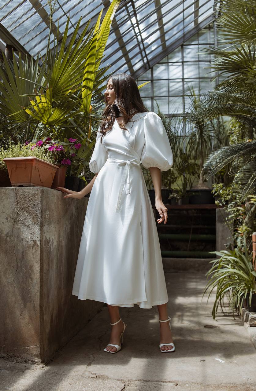 Woman in a white dress standing in a greenhouse filled with plants.