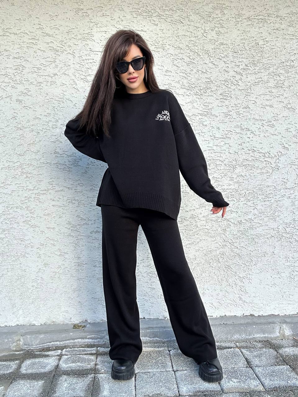 Woman wearing a black outfit with a logo against a textured wall.