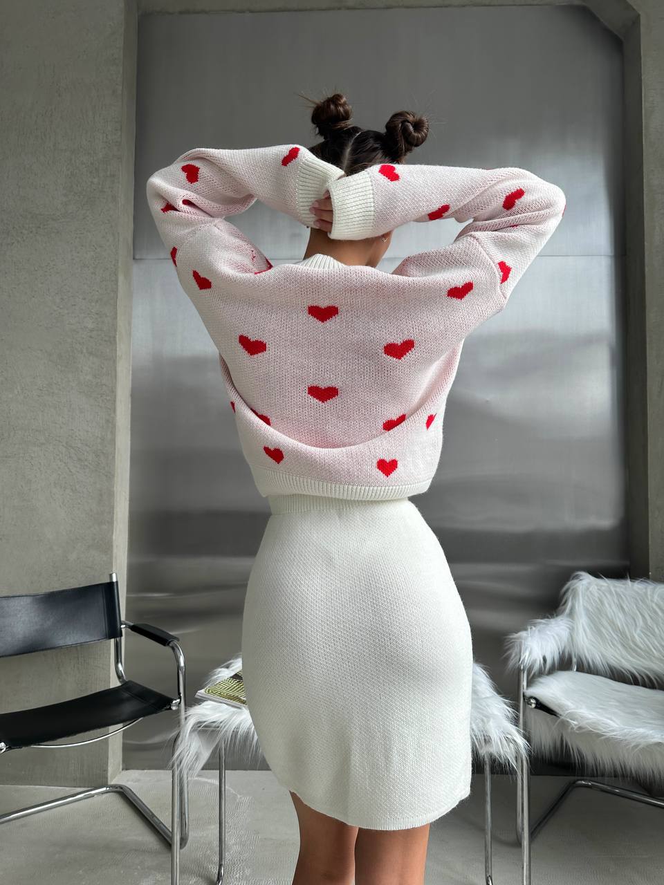 Person wearing a pink sweater with red hearts and white skirt in a room.